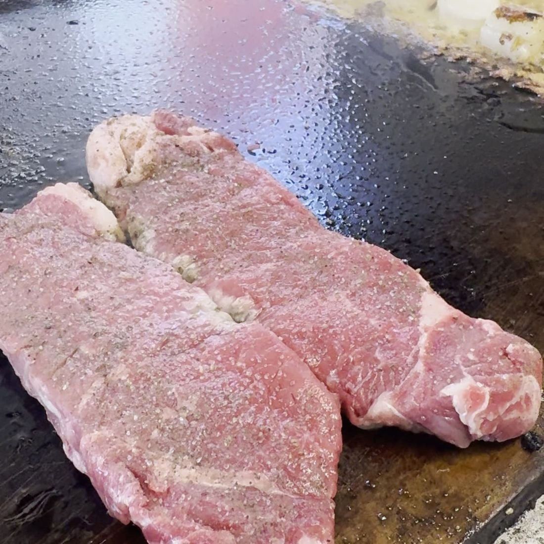 Two New York strip steaks seasoned and searing on the flat-top, steam rising